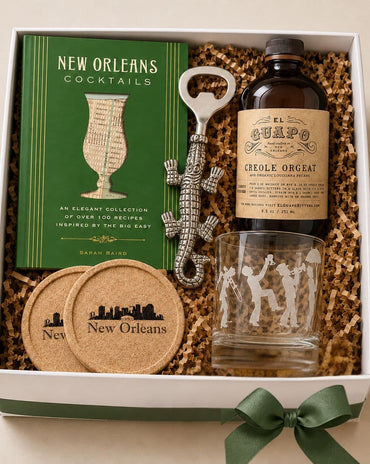 A New Orleans-themed gift box set containing a green 'New Orleans Cocktails' book, an El Guapo bottle of Creole Orgat, a silver alligator-shaped bottle opener, a clear etched tumbler, and two cork coasters inside a white gift box with shredded paper.