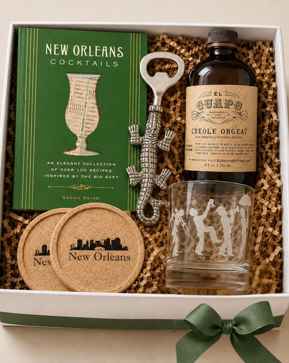 A New Orleans-themed gift box set containing a green 'New Orleans Cocktails' book, an El Guapo bottle of Creole Orgat, a silver alligator-shaped bottle opener, a clear etched tumbler, and two cork coasters inside a white gift box with shredded paper.