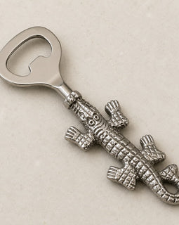 A silver-toned metal bottle opener shaped like a detailed alligator, displayed diagonally on a light beige textured background.