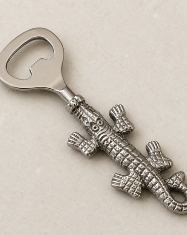 A silver-toned metal bottle opener shaped like a detailed alligator, displayed diagonally on a light beige textured background.