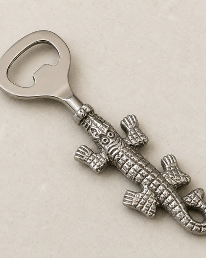A silver-toned metal bottle opener shaped like a detailed alligator, displayed diagonally on a light beige textured background.