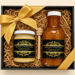 A honey gift set in a black box with shredded brown paper filler, a large gold ribbon bow, and two labeled jars: a small jar of honey and a larger bottle of honey, plus a wooden honey dipper. The labels read 'Anceley's Honey Farm' and 'Carencho, LA'.