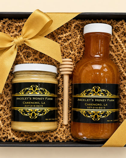 A honey gift set in a black box with shredded brown paper filler, a large gold ribbon bow, and two labeled jars: a small jar of honey and a larger bottle of honey, plus a wooden honey dipper. The labels read 'Anceley's Honey Farm' and 'Carencho, LA'.