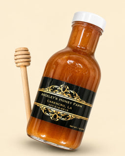 A bottle of amber honey with a black and gold label reading 'Ancelet's Honey Farm Carencro, LA' next to a wooden honey dipper on a light beige background.