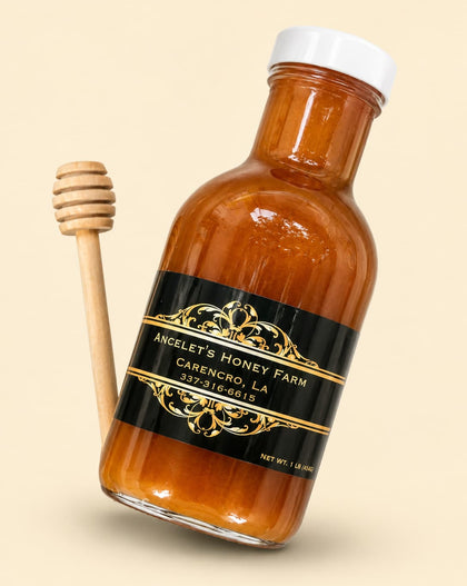 A bottle of amber honey with a black and gold label reading 'Ancelet's Honey Farm Carencro, LA' next to a wooden honey dipper on a light beige background.