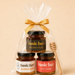 A gift set of three jars of Basic Bee honey in a clear cellophane bag tied with a gold ribbon, with a wooden honey dipper on the right, against a solid beige background.