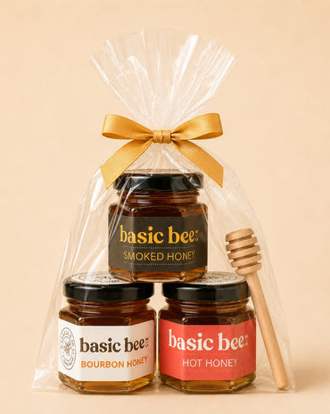 A gift set of three jars of Basic Bee honey in a clear cellophane bag tied with a gold ribbon, with a wooden honey dipper on the right, against a solid beige background.