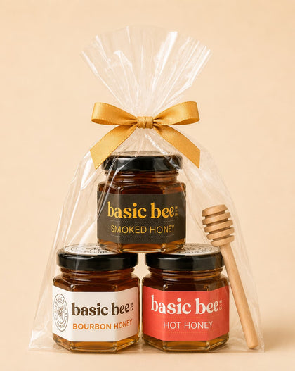 A gift set of three jars of Basic Bee honey in a clear cellophane bag tied with a gold ribbon, with a wooden honey dipper on the right, against a solid beige background.