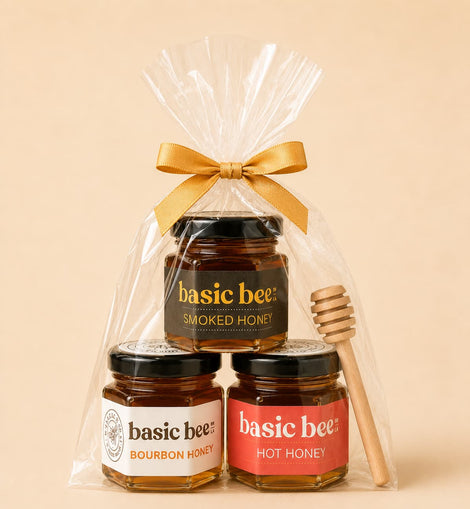 A gift set of three jars of Basic Bee honey in a clear cellophane bag tied with a gold ribbon, with a wooden honey dipper on the right, against a solid beige background.