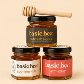 Three glass jars of Basic Bee honey arranged in a pyramid on a warm beige background. The top jar is Smoked Honey with a black label and a wooden honey dipper resting on its lid.