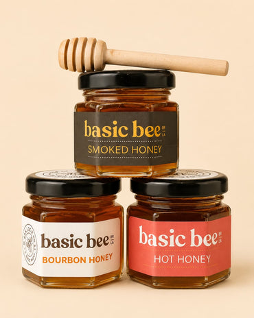 Three glass jars of Basic Bee honey arranged in a pyramid on a warm beige background. The top jar is Smoked Honey with a black label and a wooden honey dipper resting on its lid.