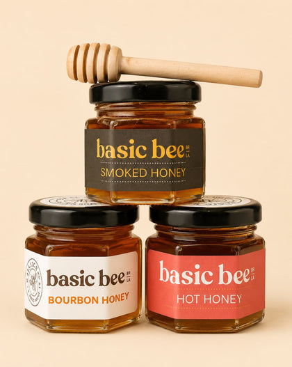 Three glass jars of Basic Bee honey arranged in a pyramid on a warm beige background. The top jar is Smoked Honey with a black label and a wooden honey dipper resting on its lid.