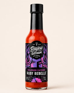 A bottle of Bayou Gotham Ruby Rebel hot sauce on a solid light beige background. The bottle is filled with bright red-orange sauce and features a black cap and a black label with purple and pink graphic details.