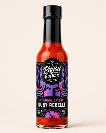 A bottle of Bayou Gotham Ruby Rebel hot sauce on a solid light beige background. The bottle is filled with bright red-orange sauce and features a black cap and a black label with purple and pink graphic details.