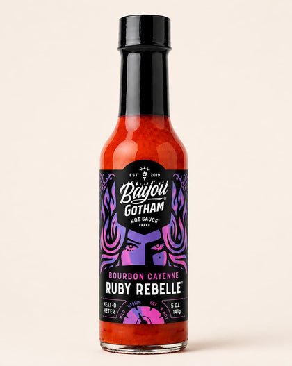 A bottle of Bayou Gotham Ruby Rebel hot sauce on a solid light beige background. The bottle is filled with bright red-orange sauce and features a black cap and a black label with purple and pink graphic details.