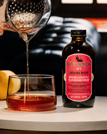 A bottle of BitterMilk No. 4 cocktail mixer stands on a tabletop next to a glass filled with amber liquid, with a strainer pouring liquid into the glass and a lemon nearby.