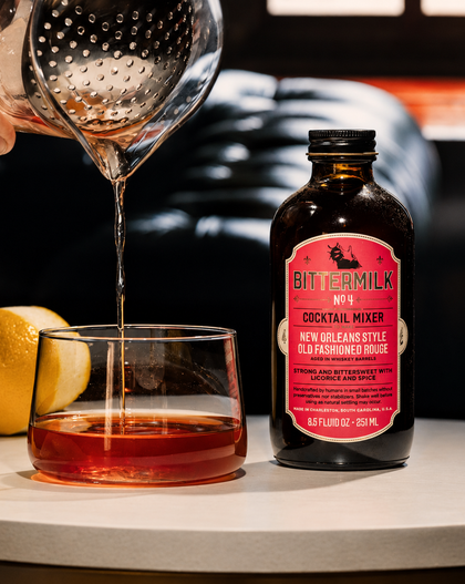 A bottle of BitterMilk No. 4 cocktail mixer stands on a tabletop next to a glass filled with amber liquid, with a strainer pouring liquid into the glass and a lemon nearby.
