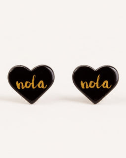 A pair of heart-shaped black enamel stud earrings with the word 'nola' written in gold cursive script, displayed on a clean off-white background.