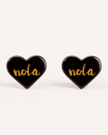 A pair of heart-shaped black enamel stud earrings with the word 'nola' written in gold cursive script, displayed on a clean off-white background.