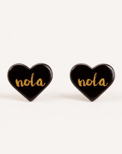 A pair of heart-shaped black enamel stud earrings with the word 'nola' written in gold cursive script, displayed on a clean off-white background.