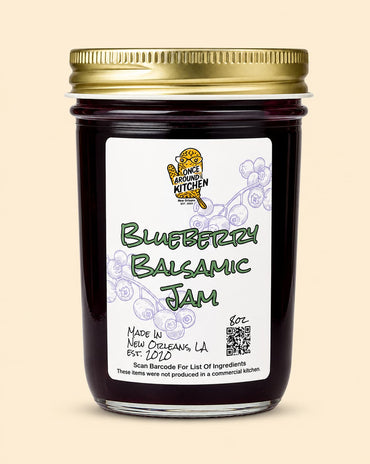 A glass jar of Blueberry Balsamic Jam with a gold metal lid and a white label featuring the 'Once Loving Kitchen' logo, set against a solid beige background.