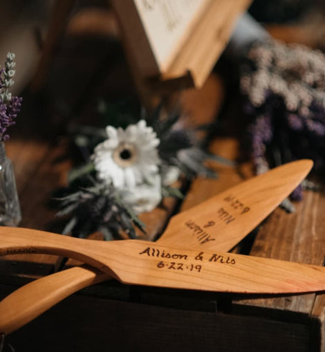 Cherry wood cake knife and server set with engraved names displayed on a rustic table with flowers.