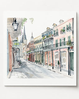 A square watercolor painting of a European street scene with pastel-colored buildings, balconies, shop fronts, and a bicycle on a sidewalk, shown against a light gray background.
