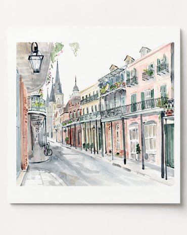 A square watercolor painting of a European street scene with pastel-colored buildings, balconies, shop fronts, and a bicycle on a sidewalk, shown against a light gray background.