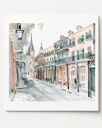 A square watercolor painting of a European street scene with pastel-colored buildings, balconies, shop fronts, and a bicycle on a sidewalk, shown against a light gray background.