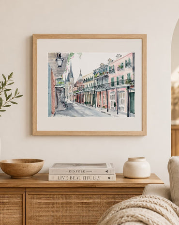 A framed watercolor painting of a historic street scene with colorful buildings hangs on a cream wall above a wooden console table styled with books, vases, and a bowl.