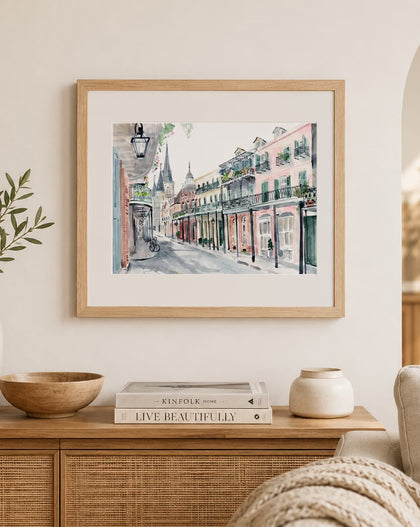 A framed watercolor painting of a historic street scene with colorful buildings hangs on a cream wall above a wooden console table styled with books, vases, and a bowl.