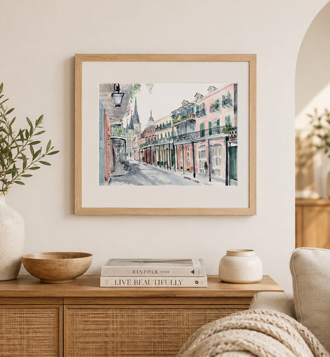 A framed watercolor painting of a historic street scene with colorful buildings hangs on a cream wall above a wooden console table styled with books, vases, and a bowl.