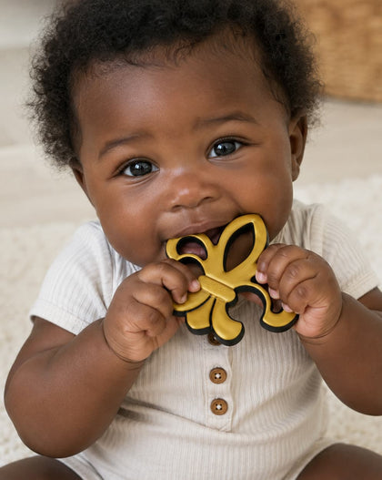 A baby wearing a cream ribbed short-sleeve romper with brown buttons, holding a yellow and black Saints teether in their mouth while sitting on a soft cream rug.