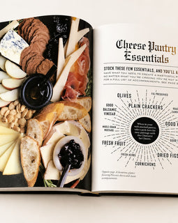 An open hardcover book showing a cheese and charcuterie spread on one page and a 'Cheese Pantry Essentials' infographic on the other, photographed from above on a light neutral background.