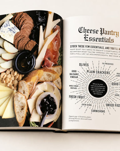 An open hardcover book showing a cheese and charcuterie spread on one page and a 'Cheese Pantry Essentials' infographic on the other, photographed from above on a light neutral background.