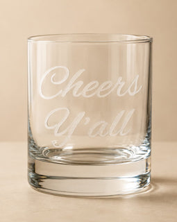 A clear glass rocks tumbler with the phrase 'Cheers Y'all' etched in white script on the front, set against a warm beige background.