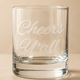 A clear glass rocks tumbler with the phrase 'Cheers Y'all' etched in white script on the front, set against a warm beige background.