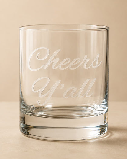 A clear glass rocks tumbler with the phrase 'Cheers Y'all' etched in white script on the front, set against a warm beige background.