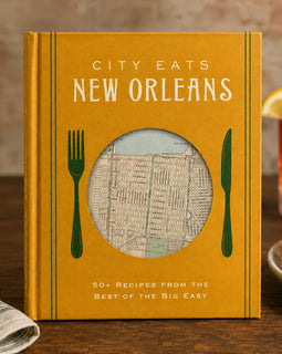 A hardcover cookbook titled "City Eats New Orleans" stands on a wooden table in a restaurant-like setting. The cover is mustard yellow with white title text, green fork and knife illustrations, and a circular window showing a vintage street map.