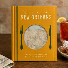 A hardcover cookbook titled "City Eats New Orleans" stands on a wooden table in a restaurant-like setting. The cover is mustard yellow with white title text, green fork and knife illustrations, and a circular window showing a vintage street map.