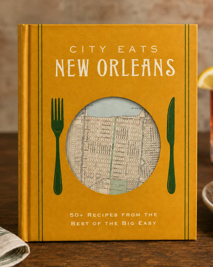 A hardcover cookbook titled "City Eats New Orleans" stands on a wooden table in a restaurant-like setting. The cover is mustard yellow with white title text, green fork and knife illustrations, and a circular window showing a vintage street map.