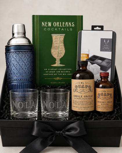 A curated cocktail gift set arranged in a black box with a black ribbon. The set includes a blue patterned cocktail shaker, two clear engraved glasses reading 'NOLA', a green book titled 'New Orleans Cocktails', two amber bottles labeled 'El Campo', and a boxed ice tray set.