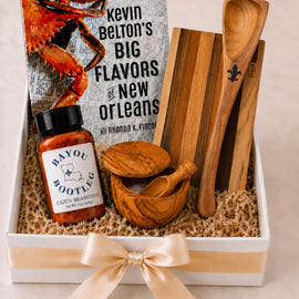 A gift box set featuring Kevin Belton's book 'Big Flavors of New Orleans,' a bottle of Bayou Bootleg Cajun Brisket sauce, wooden utensils, and wooden truffle-shaped items arranged in a white box with a cream ribbon.