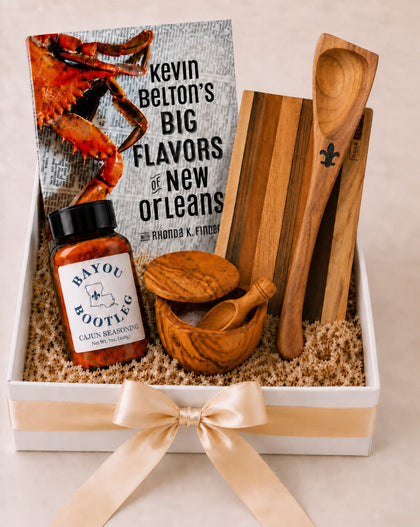 A gift box set featuring Kevin Belton's book 'Big Flavors of New Orleans,' a bottle of Bayou Bootleg Cajun Brisket sauce, wooden utensils, and wooden truffle-shaped items arranged in a white box with a cream ribbon.