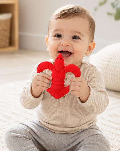 A baby sitting on a light-colored rug holds a bright red CRAWFISH-shaped teething toy. The baby is wearing a beige long-sleeve top and light gray pants in a softly lit, cozy room.