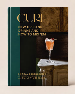 The book 'Cure: New Orleans Drinks and How to Mix 'Em' by Neal Bodenheimer and Emily Timberlake, featuring a dark teal and black cover with gold lettering and a photograph of an orange cocktail on a marble table.