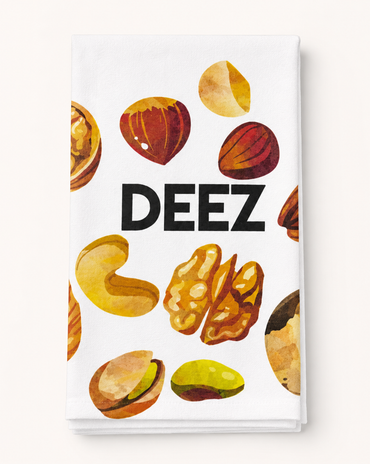 A white kitchen towel featuring a watercolor-style print of various nuts and the word 'DEEZ' in bold black letters, shown against a light beige background.