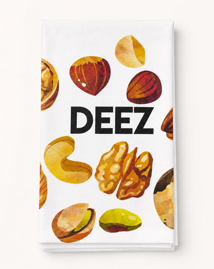 A white kitchen towel featuring a watercolor-style print of various nuts and the word 'DEEZ' in bold black letters, shown against a light beige background.