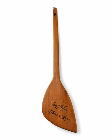 A single wooden paddle with a long handle and broad blade, shown upright on a plain white background. The blade is engraved with the phrase 'First You, Make a Roux".