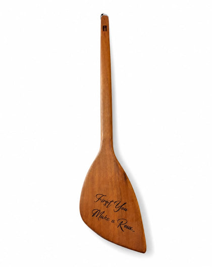 A single wooden paddle with a long handle and broad blade, shown upright on a plain white background. The blade is engraved with the phrase 'First You, Make a Roux".